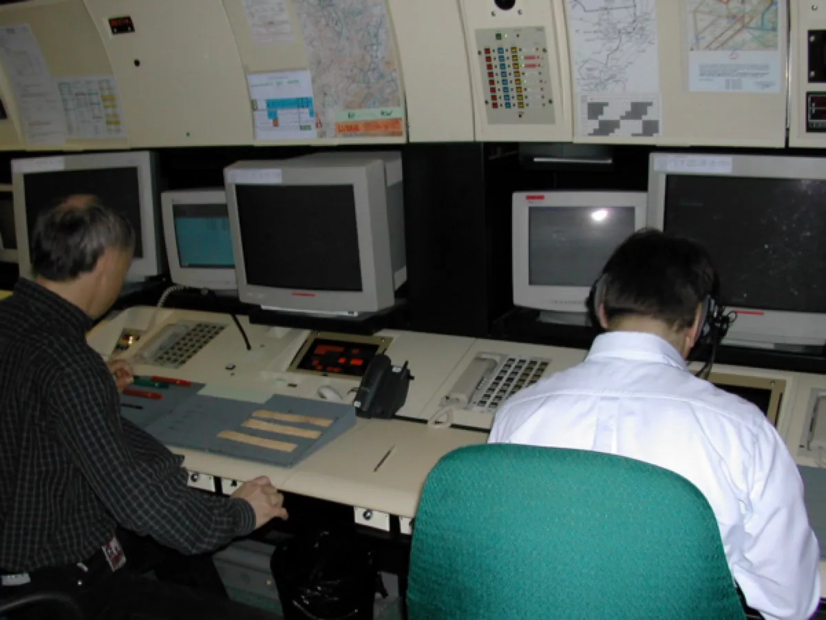 An air traffic control approach room. Controllers sit at large radar screens in a dim room of 1990s equipment, watching shared displays.