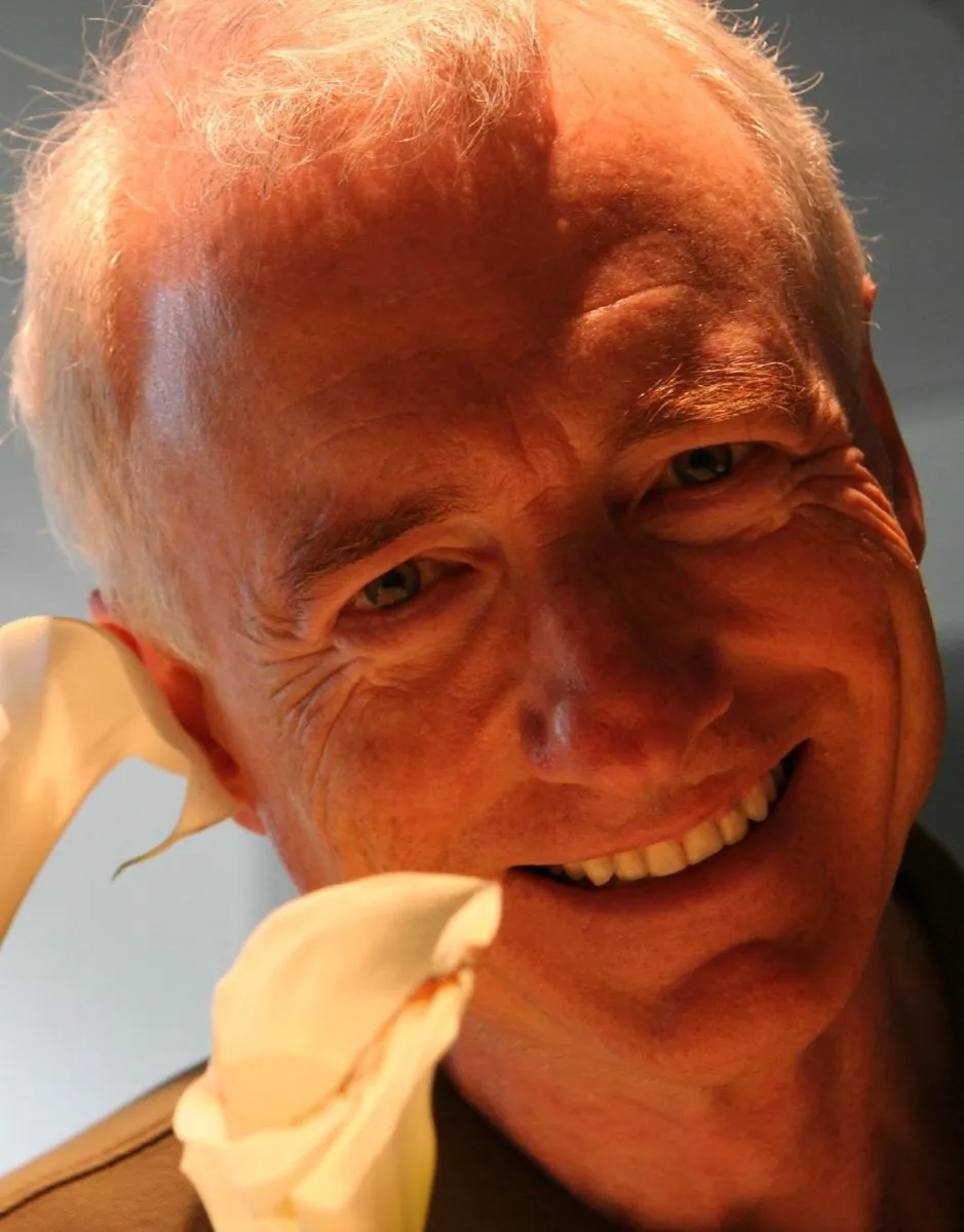 A smiling man with glasses and grey hair, photographed close up against a plain background.