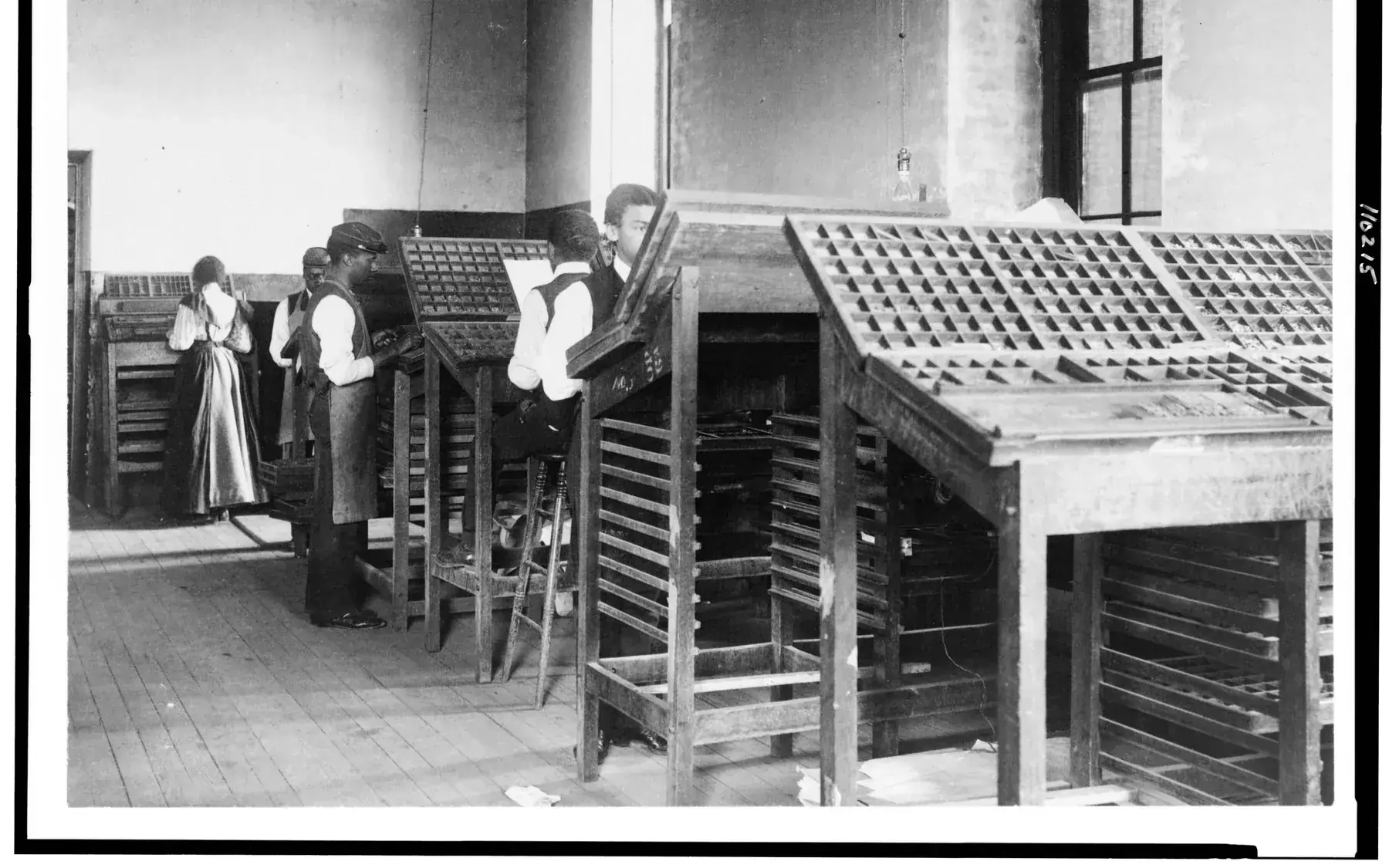Black-and-white 1899 photograph of several men in a print shop setting metal type by hand at composing frames.
