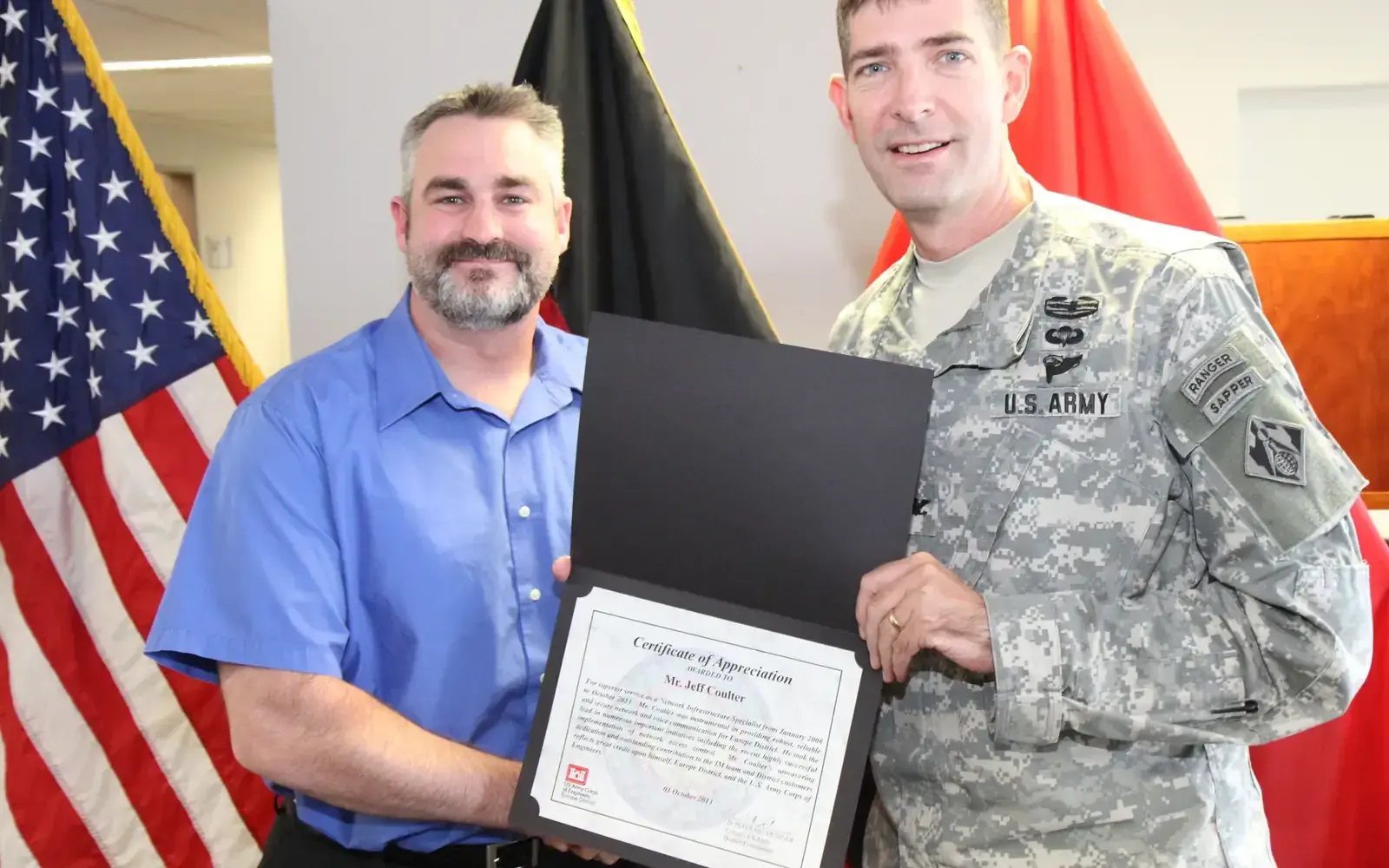 A district commander presents a certificate of appreciation to a network infrastructure specialist during an office award ceremony.